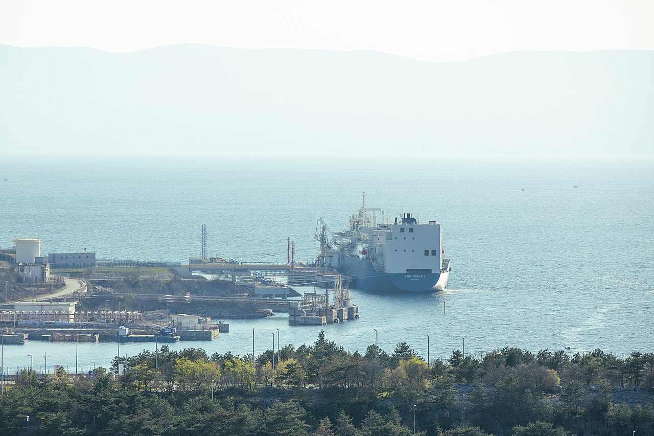A large oil tanker docked at a port with pipelines and storage facilities visible alongside