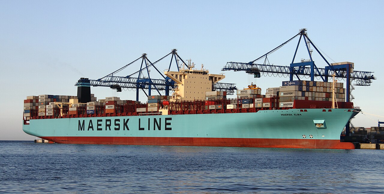 A Maersk Line container ship loaded with shipping containers at a port terminal, with blue container cranes in the background.