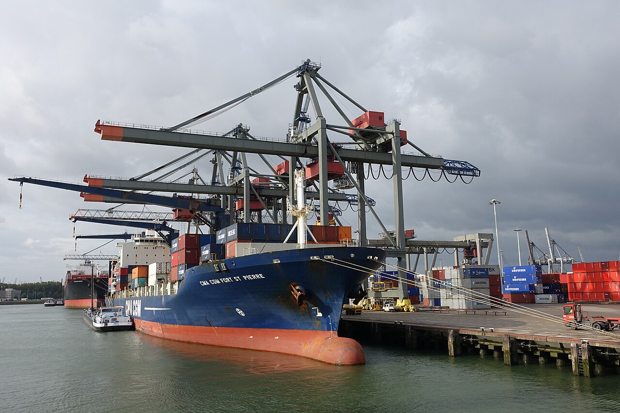 A large container ship docked at a port terminal with massive container cranes towering overhead, surrounded by stacked shipping containers.